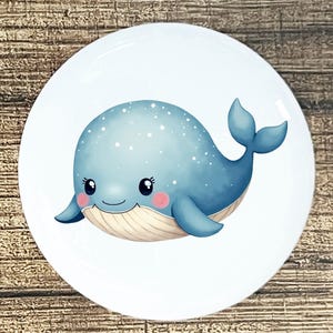 May include: A white, round plate featuring a cartoon whale illustration. The whale is light blue with white star accents, rosy cheeks, and a smiling expression. The plate has a glossy finish and is set against a wooden background.
