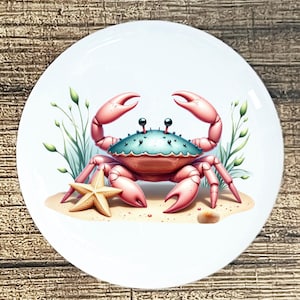 May include: White ceramic plate featuring a cartoon crab with pink claws and a teal shell. A starfish and seashell are on the sand. Green seaweed is in the background. The plate has a glossy finish and is suitable for serving food.