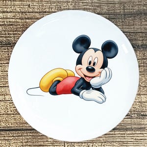 May include: White ceramic plate featuring a colorful illustration of Mickey Mouse. Mickey is depicted in a relaxed pose, wearing red shorts, yellow shoes, and white gloves. The plate has a glossy finish and a round shape.
