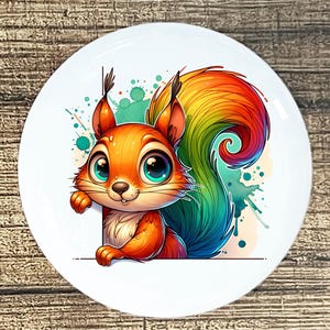 May include: A white ceramic plate featuring a cartoon squirrel with orange fur, large blue eyes, and a rainbow-colored tail. The squirrel is holding a blank white sign, with teal and green watercolor splashes in the background.