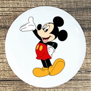 May include: A white, round plate featuring a cartoon image of Mickey Mouse. Mickey is depicted in his classic attire: red shorts with yellow buttons, yellow shoes, white gloves, and a black body and ears. He is smiling and waving.