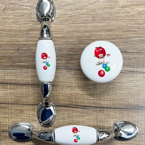 May include: Three white ceramic cabinet knobs with silver metal accents. The knobs feature a cartoon M&M's character with red, green, blue, and yellow M&M's.