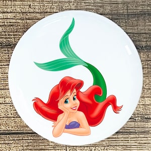 May include: A white round sticker with a cartoon image of Ariel, the Little Mermaid, with red hair, a purple seashell bra, and a green tail.
