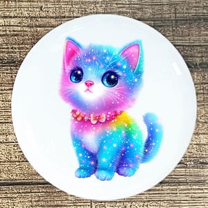 May include: A colorful illustration of a kitten with a rainbow-colored body and sparkling details. The kitten has large blue eyes, a pink nose, and a pink collar with a bow. The image is on a white, round surface.