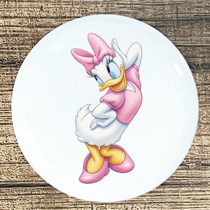 May include: A white ceramic plate featuring a cartoon image of Daisy Duck. Daisy is depicted in a pink dress, shoes, and bow, with a yellow beak and blue eyes. The plate has a glossy finish and is set against a wooden background.