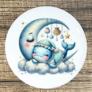 May include: A white ceramic plate featuring a whimsical illustration of a sleeping blue whale wearing a nightcap and pajamas, resting on a cloud in front of a crescent moon. Decorative elements include hanging seashells, stars, and a soft, dreamy aesthetic.