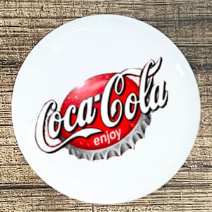 May include: A white round button with a red and white Coca-Cola logo. The logo is a stylized bottle cap with the words "Coca-Cola" in white and "enjoy" in black.