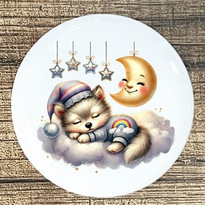 May include: A white ceramic plate featuring a sleeping wolf pup wearing a nightcap and sweater with a rainbow design. A smiling crescent moon and hanging stars complete the whimsical design. The plate has a diameter of approximately 8 inches.