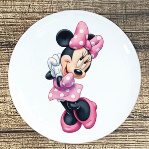 May include: White ceramic plate featuring a cartoon illustration of Minnie Mouse. Minnie is depicted in a pink polka-dot dress, matching bow, and shoes. The plate has a glossy finish and is set against a wooden background.