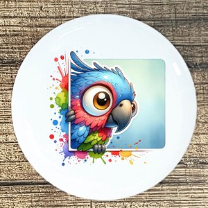 May include: White ceramic plate featuring a colorful cartoon parrot design. The parrot has blue, red, and green feathers, with a large brown eye and a gray beak. The design is framed by a square and surrounded by colorful paint splatters.