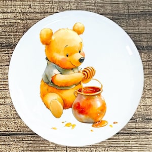 May include: A white ceramic plate featuring a watercolor illustration of a yellow bear with a honey pot. The bear is wearing a gray shirt and holding a honey dipper, with honey dripping into the pot. The plate has a glossy finish.