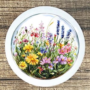 May include: A round, white-rimmed decorative plate featuring a vibrant watercolor painting of a wildflower meadow. The artwork showcases a variety of colorful flowers, including yellow daisies, pink blossoms, and purple lavender, set against a backdrop of green foliage.