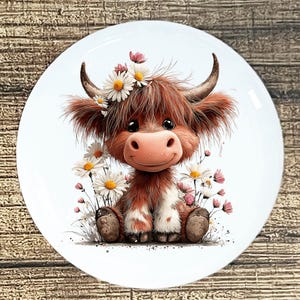 May include: A white ceramic plate featuring a cartoon illustration of a Highland cow. The cow has brown and white fur, a pink nose, and is adorned with a daisy flower crown. Daisies and pink flowers surround the cow.