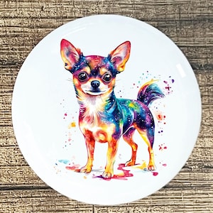 May include: A round, white ceramic plate featuring a colorful watercolor illustration of a Chihuahua dog. The dog's fur is a vibrant mix of blues, purples, oranges, and yellows, with a starry galaxy effect. The plate has a glossy finish.