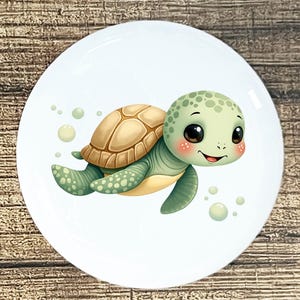 May include: A white ceramic plate featuring a cartoon sea turtle illustration. The turtle has a light green head and limbs, a tan shell, and rosy cheeks. Bubbles surround the turtle, and the plate has a glossy finish.