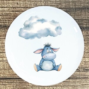 May include: A white ceramic plate featuring a watercolor illustration of a blue donkey character sitting below a fluffy blue cloud. The donkey has large ears and a friendly expression. The plate has a glossy finish.
