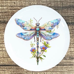 May include: A white ceramic plate featuring a colorful dragonfly design. The dragonfly has blue, purple, and green wings with a mosaic pattern. Below the dragonfly are purple flowers with orange centers and green leaves.