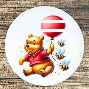 May include: A white ceramic plate featuring a cartoon bear in a red shirt holding a red balloon with white stripes. Three cartoon bees with blue wings fly nearby. The plate has a glossy finish.