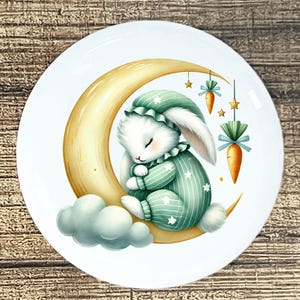 May include: White ceramic plate featuring a sleeping bunny in green pajamas and cap, resting on a golden crescent moon. The design includes a cloud, carrots, and stars, creating a whimsical, dreamy aesthetic.