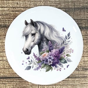 May include: White ceramic plate featuring a watercolor illustration of a horse's head with a flowing mane. The horse is surrounded by purple flowers, green leaves, and butterflies. The plate has a glossy finish and is set against a wooden background.