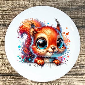 May include: A white ceramic plate featuring a cartoon squirrel with large blue eyes and orange fur. The squirrel is peeking over a white surface, with colorful paint splatters around it. The plate has a glossy finish.