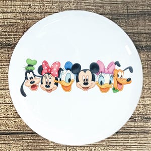 May include: White ceramic plate featuring a colorful illustration of Disney characters including Goofy, Minnie Mouse, Donald Duck, Mickey Mouse, Daisy Duck, and Pluto. The plate has a glossy finish and is suitable for serving food.