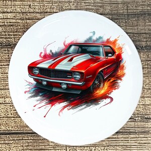 May include: A white ceramic plate featuring a vibrant illustration of a classic red sports car with white racing stripes. The car is depicted with fiery accents and a dynamic, artistic style, evoking speed and power.