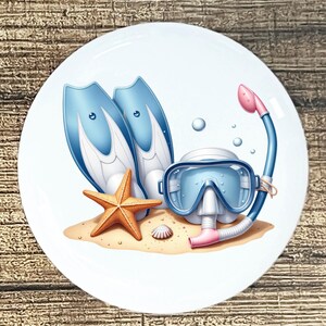 May include: A white ceramic plate featuring a beach scene with blue and white flippers, a diving mask, and a snorkel. An orange starfish and a small seashell rest on the sand. Bubbles float in the background, evoking a summer theme.