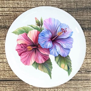 May include: White ceramic plate featuring a detailed floral design. Two hibiscus flowers, one pink and the other blue and purple, are the focal point. Green leaves and a bud complete the botanical illustration.