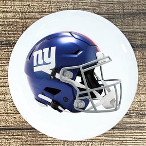 May include: A blue football helmet with the letters "ny" in white on the side. The helmet has a gray face mask and a red stripe on top. The helmet is on a white plate.