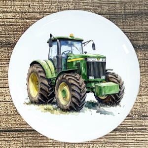May include: White ceramic plate featuring a watercolor illustration of a green tractor with large tires. The tractor has yellow accents and is set against a white background. The plate is round and has a glossy finish.