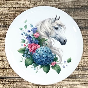 May include: White ceramic plate featuring a detailed illustration of a white horse's head with flowing mane, surrounded by blue hydrangeas, pink roses, and green foliage. The design is set against a white background.