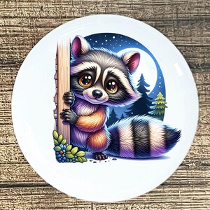 May include: A white ceramic plate featuring a cartoon raccoon with big eyes, peeking from behind a tree trunk. The background shows a night scene with a full moon, stars, and pine trees. The raccoon has a black and gray mask and striped tail.