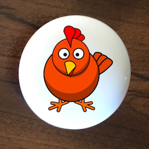 May include: A white ceramic knob with a cartoon image of a brown chicken with red comb and wattle. The chicken has large, round eyes and a yellow beak.