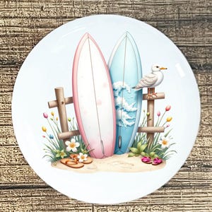 May include: A decorative plate featuring a beach scene with two surfboards, one pink and one blue, leaning against a wooden fence. A seagull perches on the fence, and sandals and flowers are scattered on the sand. The plate has a white background.