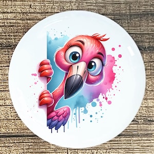 May include: A white ceramic plate featuring a cartoon flamingo with pink feathers, a black beak, and large blue eyes. The flamingo peeks out from behind a white surface against a watercolor background of pink and blue.