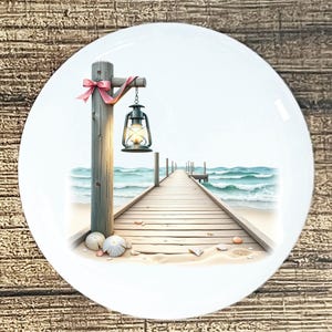 May include: White ceramic plate featuring a coastal scene. A wooden pier extends into turquoise water under a bright sky. A lantern hangs from a wooden post adorned with a pink ribbon. Seashells and sand are at the base of the post.