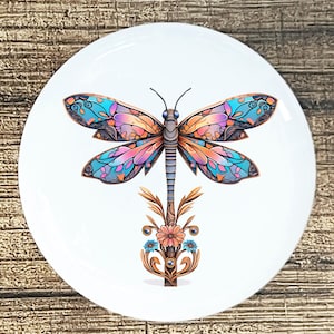 May include: A white ceramic plate featuring a colorful dragonfly design. The dragonfly has intricate details with blue, pink, and orange wings. The design includes a floral element at the base, adding a decorative touch.