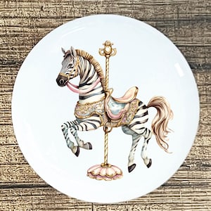 May include: A white ceramic plate featuring a carousel zebra illustration. The zebra has black and white stripes, a pink saddle, and a gold pole. The design is detailed with gold accents and a pink base.