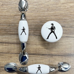 May include: Three ceramic cabinet knobs with a black and white silhouette of a person playing a guitar. The knobs are white with silver metal accents.