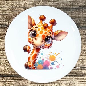 May include: A white ceramic plate featuring a cartoon giraffe with orange spots and big blue eyes. The giraffe is peeking from behind a white surface. Colorful paint splatters are in the background. The plate is on a wooden surface.