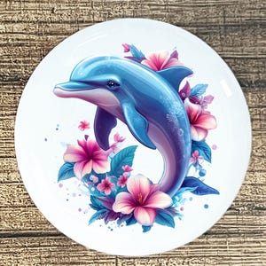 May include: A white, round plate featuring a blue dolphin leaping through pink and purple hibiscus flowers and teal leaves. The dolphin has a gradient of blue and purple hues. The plate is set against a wooden background.
