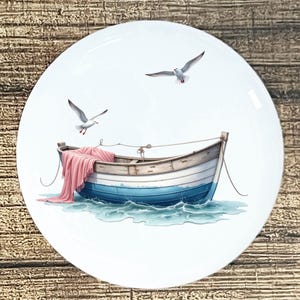 May include: A white ceramic plate featuring a watercolor illustration of a small boat on blue water with two seagulls flying above. The boat is painted in shades of blue and white, with a pink blanket draped over the side.