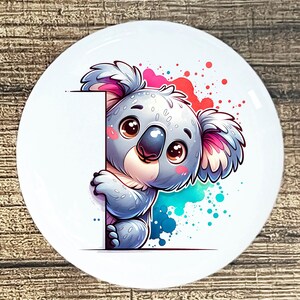 May include: A white, round decorative plate featuring a cartoon koala bear. The koala is gray with pink accents and large brown eyes, holding a blank white sign. Colorful paint splatters in red, blue, and teal surround the koala.