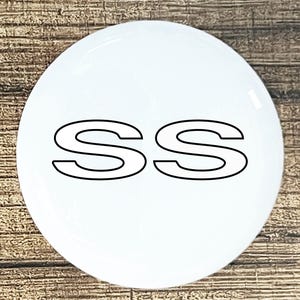 May include: A white, circular emblem with the letters "SS" in black, outlined font. The emblem is glossy and appears to be made of a smooth material. The background is a wooden surface.