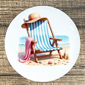 May include: A white ceramic plate featuring a beach scene. A wooden beach chair with blue and white stripes is depicted, along with a straw hat, pink towel, and sandcastle. Seashells and the ocean are also visible.