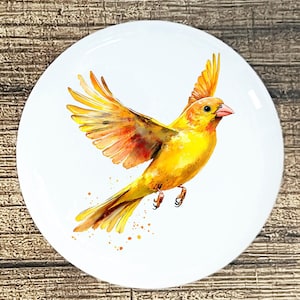 May include: A white, round decorative plate featuring a watercolor painting of a yellow bird in flight. The bird has orange and red accents on its wings and tail. Small orange paint splatters are around the bird.