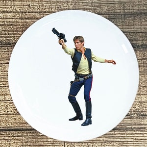 May include: White ceramic plate featuring a full-color image of a man in a yellow shirt, dark vest, and blue pants, holding a blaster. The image is a Star Wars character, Han Solo.