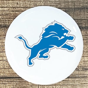 May include: A white, round coaster featuring the Detroit Lions logo. The logo is a blue lion in a leaping pose, outlined in gray, set against a white background. The coaster is on a wooden surface.