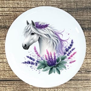 May include: A white ceramic plate featuring a watercolor illustration of a white horse's head with a lavender flower crown and flowing mane. The horse is surrounded by lavender and green foliage. The plate has a diameter of approximately 8 inches.
